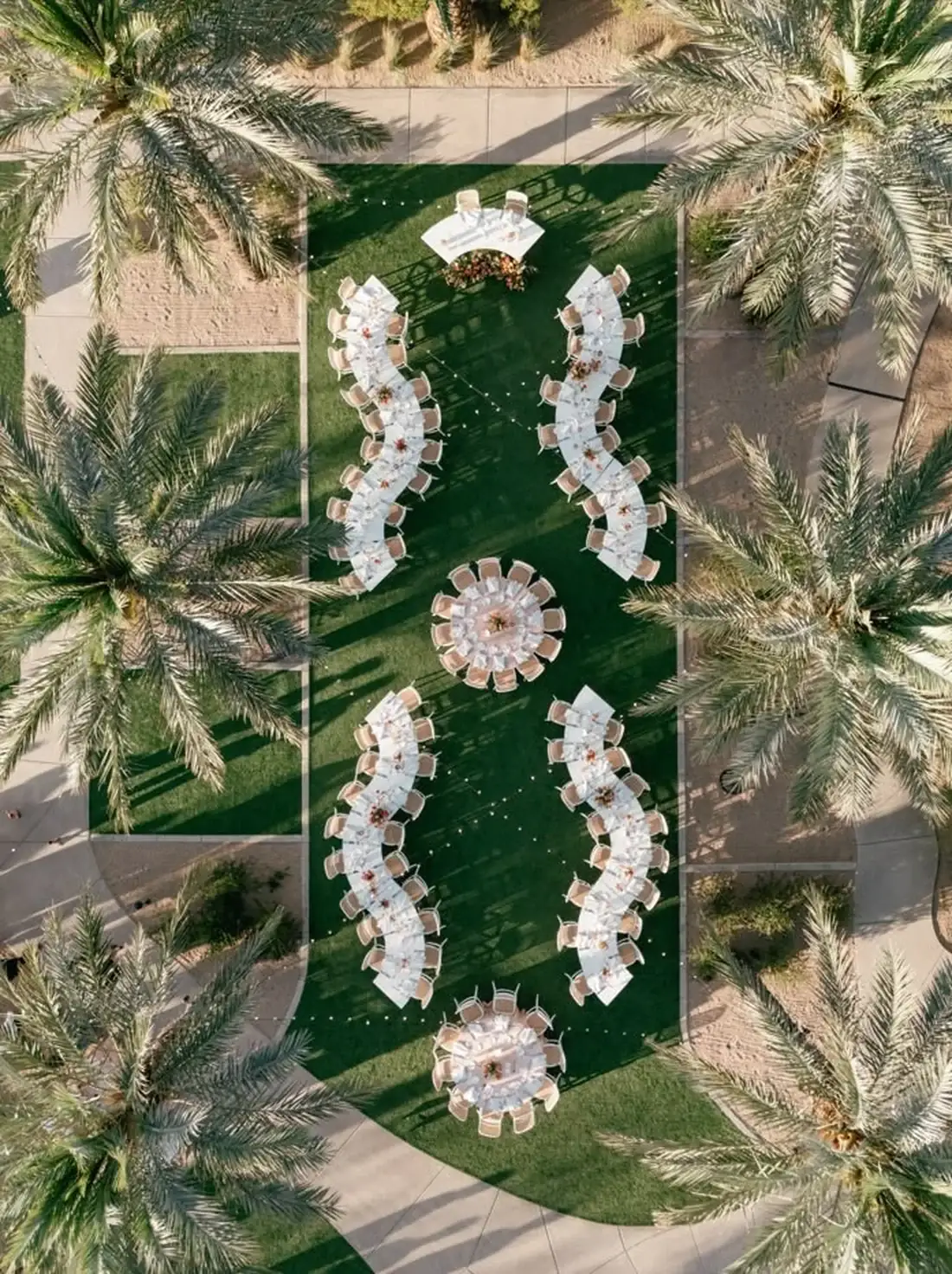 Fotografia aérea de um casamento num relvado: quatro filas de mesas serpentina em duas colunas, duas mesas redondas no eixo do meio e a mesa dos noivos no topo.